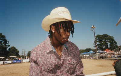 Portrait d’un jeune cowboy portant un chapeau blanc et une chemise à motifs, devant une arène de rodéo au Texas.