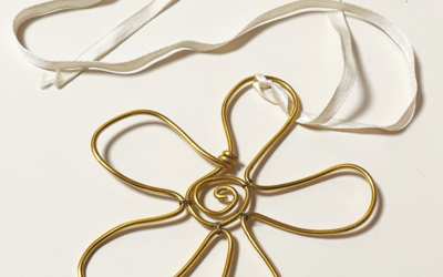 Photographie d'un collier avec un pendentif en forme de fleur fait à partir de fil d'aluminium doré, accroché à un ruban en soie blanc.