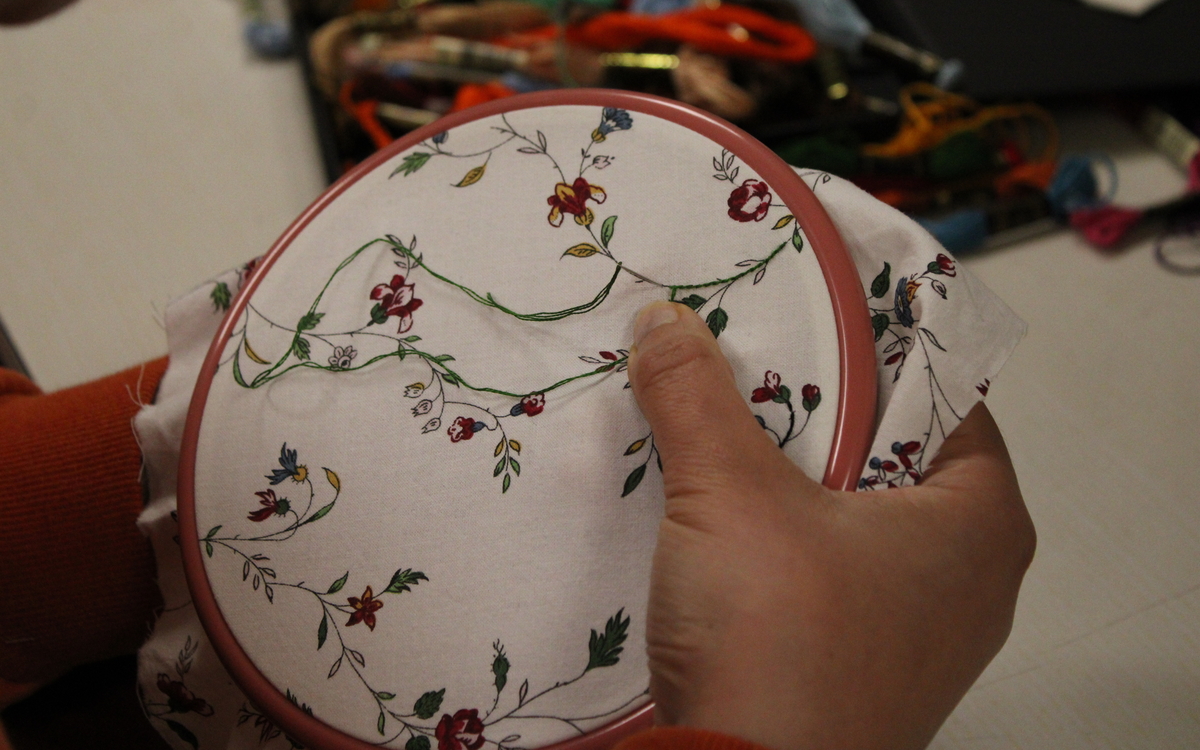 Photographie des mains d'une participante à l'atelier d'initiation à la broderie textile. Elle tient un tambour en bambou sur lequel a été tendu un tissu avec des motifs de fleurs, qu'elle vient compléter avec du fil de couleur.