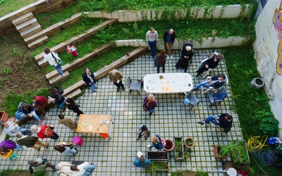 Vue aérienne d'une cour intérieure pavée où une vingtaine de personnes de tous âges sont rassemblées. Certaines sont assises autour de tables blanches et en bois, d'autres discutent debout ou sont installées sur des gradins en bois bordés de végétation. Des jardinières, des chaises et des objets personnels sont dispersés dans l'espace. Un mur à droite est orné d'une fresque colorée. L'ambiance est conviviale et détendue, évoquant un événement communautaire ou une journée portes ouvertes.