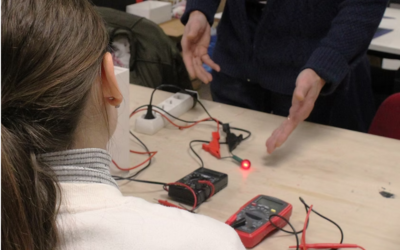 Photographie d'une participante à l'atelier brico électro vue de dos. Matériels électroniques et mains de l'animateur visibles sur la table à laquelle elle est assise.