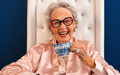 Femme senior souriante portant des lunettes, assise dans un fauteuil et tenant une tasse de thé