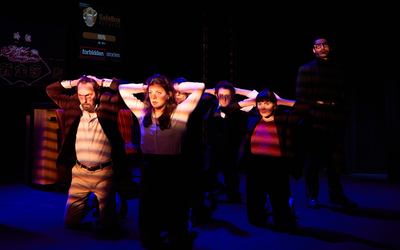 Sur une scène de théâtre, quatre personnes sont agenouillés, les mains derrière la tête