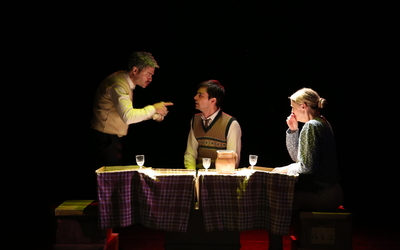 Sur une scène de théâtre, trois personnages autour d'une table 