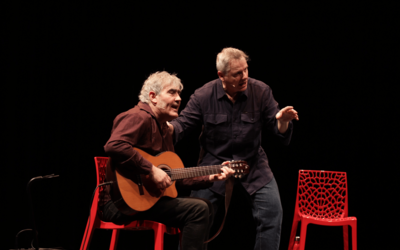 Les deux conteurs sur scène, sur fond noir, avec des chaises rouges. L'un d'eux tient une guitare