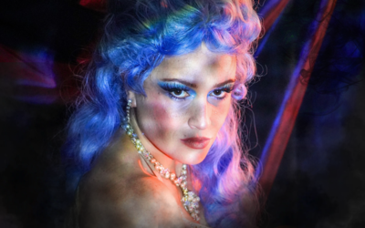 Portrait rapproché d’une artiste de cabaret aux cheveux bleu électrique et rose pastel, au maquillage drag glamour et aux faux cils marqués. Éclairée par des lumières néon bleues et roses et entourée de fumée. Une esthétique queer et spectaculaire, entre cabaret contemporain, drag et performance artistique.