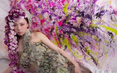 Un modèle portant des fleurs et un costume de feuilles devant un fond peint rose, violet et jaune.