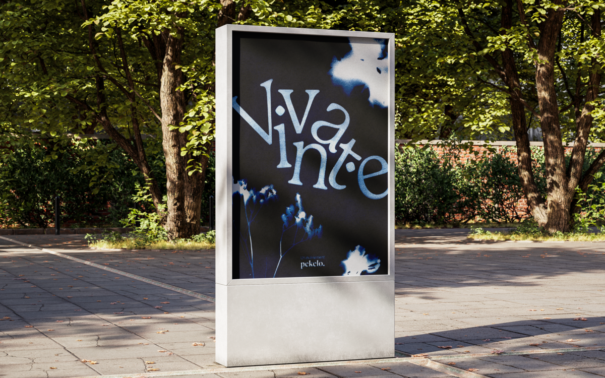 Photo d'une affiche urbaine de l'exposition Vivant.e : Au premier plan, un mobilier d'affichage urbain blanc situé sur une rue pavée déserte et ensoleillée. À l'interieur, une affiche avec comme titre "Vivant.e" et sous titre "un événement Pekelo", en arrière plan des arbres