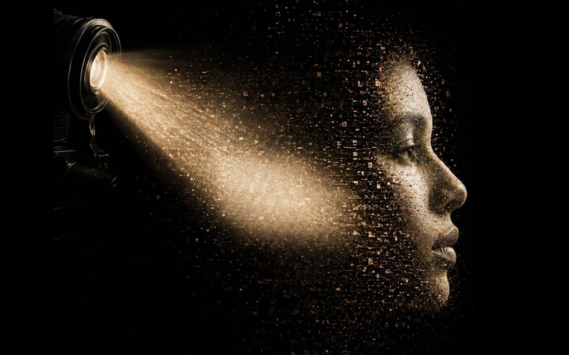 Un faisceau de lumière de projecteur éclaire un visage humain composé de particules dorées sur fond noir.